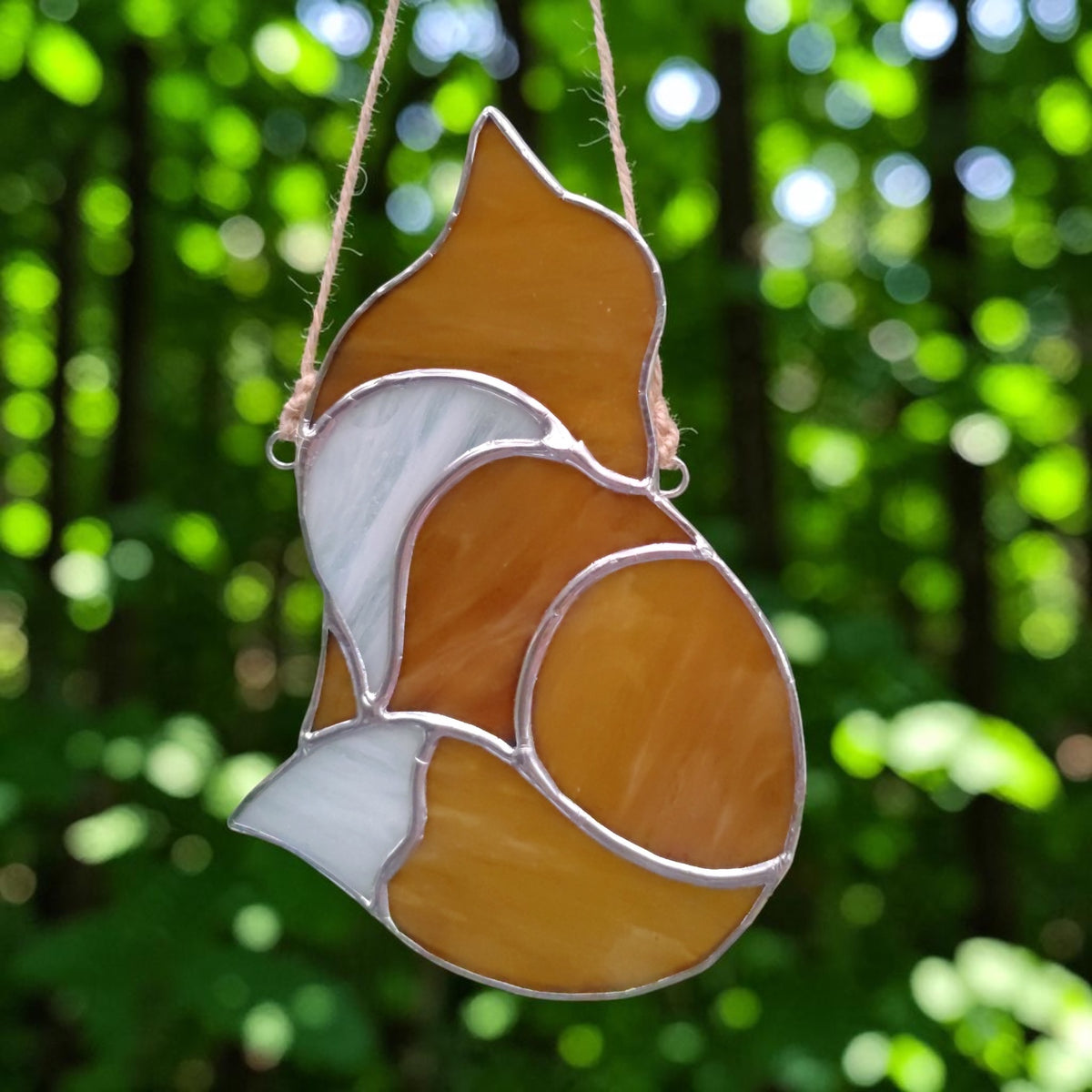 A stained glass hanging ornament shaped like a fox, featuring amber orange and white glass panels outlined in silver metal, suspended by a twine string against a blurred green forest background.