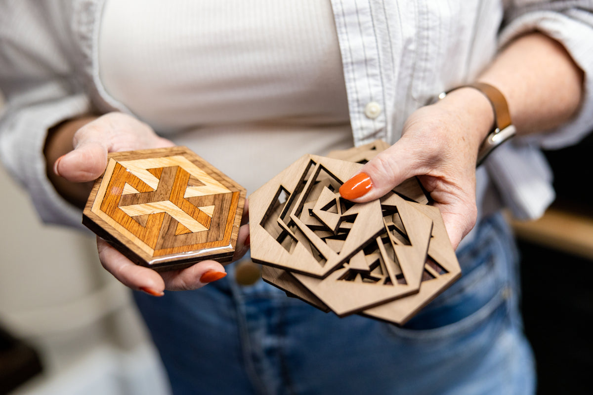 Joanna holds two hexagonal wooden coasters  in both hands. One coaster is closed, showing a geometric wood inlay design, while the other is open, revealing layered laser-cut wooden compartments.