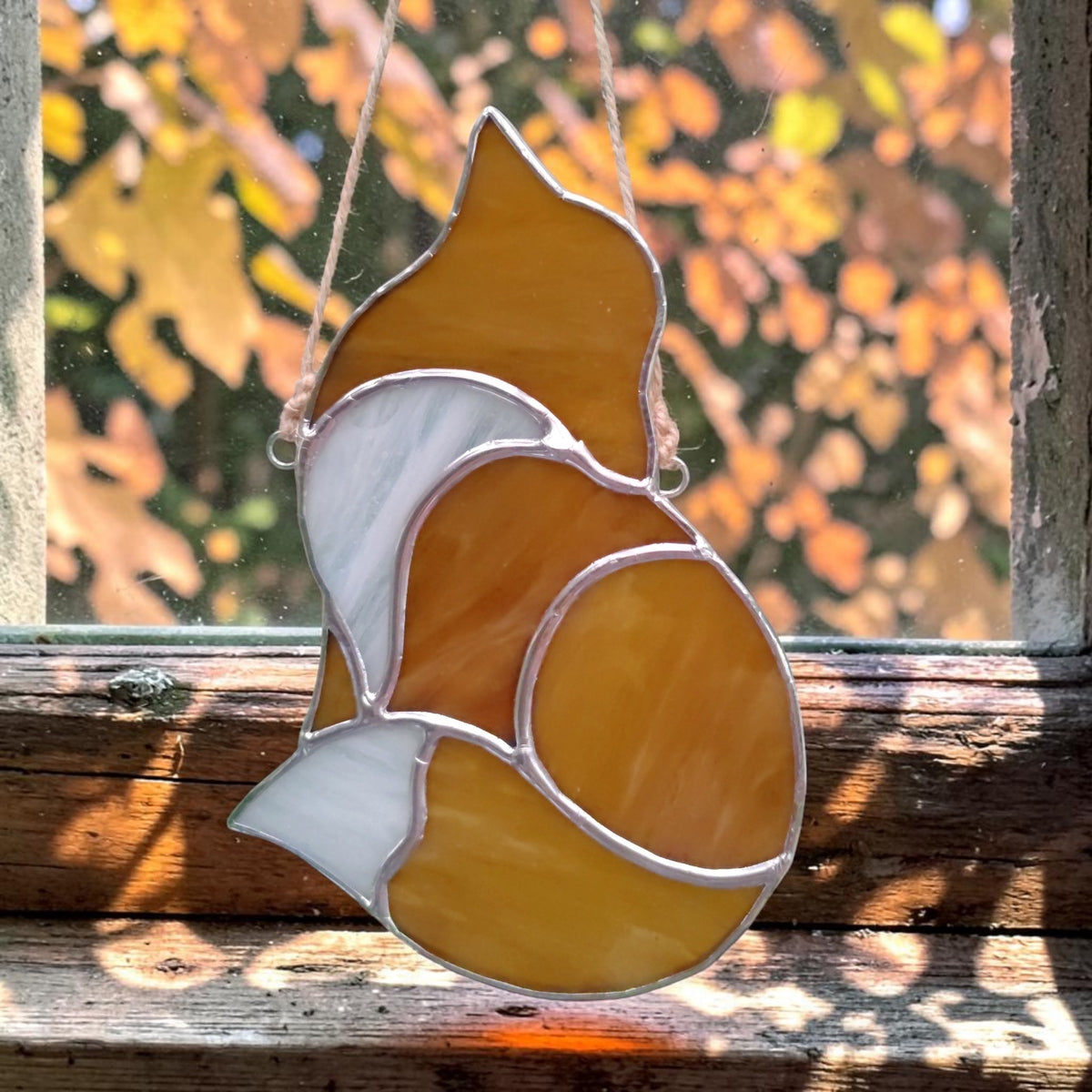 A stained glass suncatcher shaped like a fox, featuring amber yellow and white glass panels outlined in silver metal, hanging on a rustic wooden windowsill with a blurred autumn leaf background.