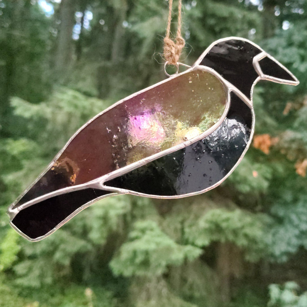 Handmade black stained-glass crow suncatcher hanging outdoors against a soft evergreen background.