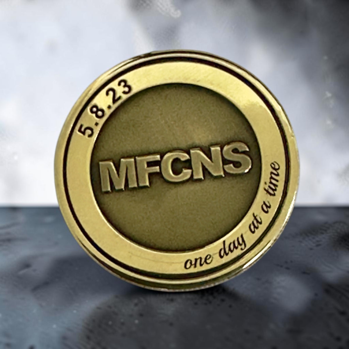 Round gold-tone commemorative coin engraved with MFCNS, the date May 8, 2023 and the phrase one day at a time