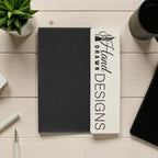 Black notebook with branding on a wooden surface with a plant and coffee cup.